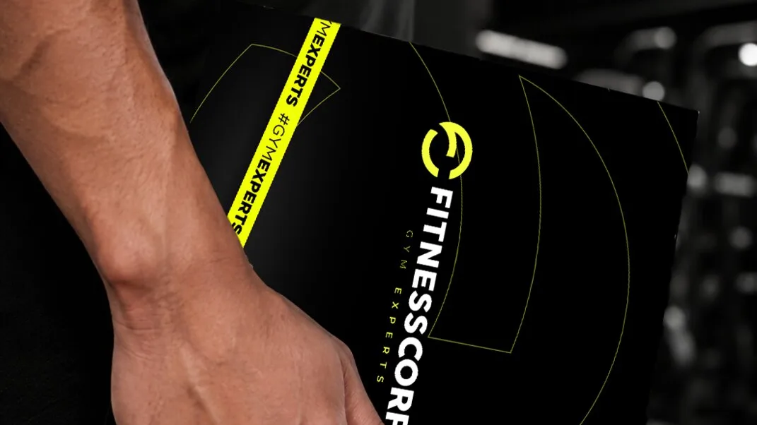 Mano sosteniendo el manual de marca negro de Fitnesscorp con la cinta #GymExperts en amarillo