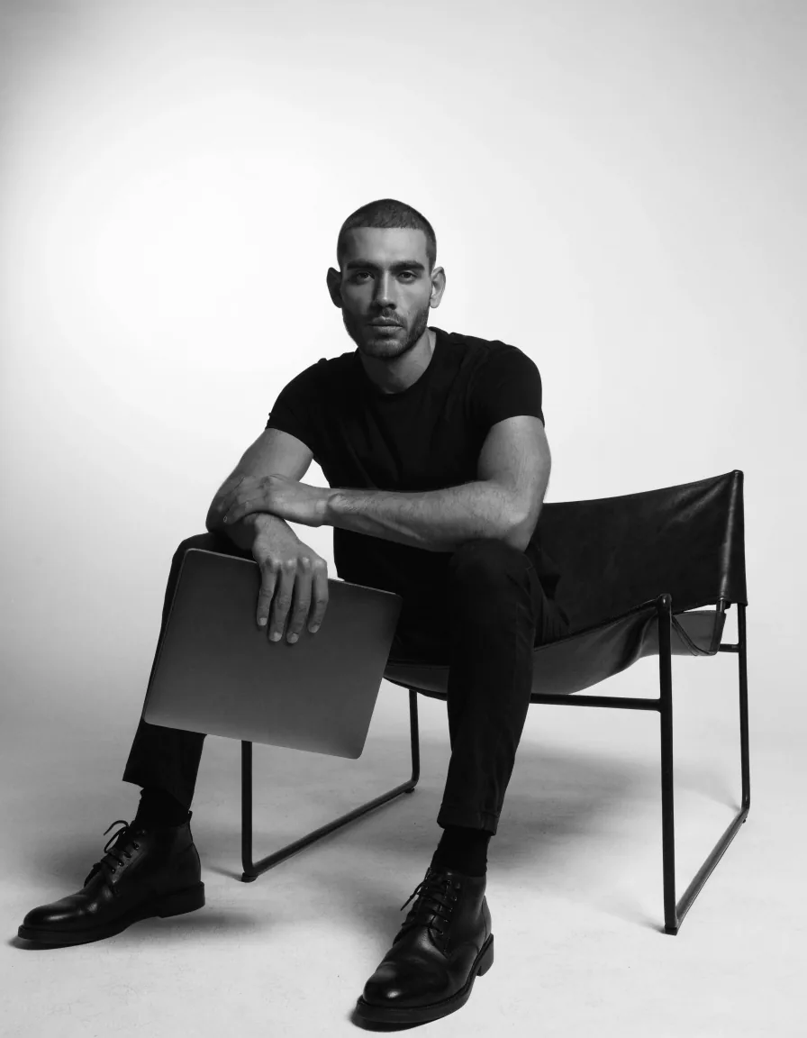 Retrato editorial en blanco y negro: un creativo sentado en una silla de piel, con una laptop bajo el brazo